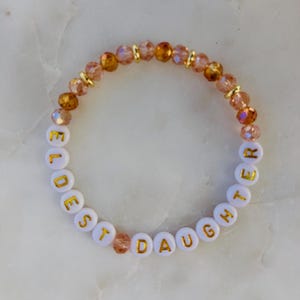 May include: A beaded bracelet with the words "ELDEST DAUGHTER" spelled out in gold letters on white beads. The bracelet features a mix of clear and amber-colored beads, with gold accents.