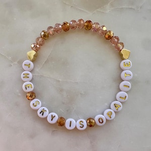 Puede incluir: Una pulsera de cuentas con las palabras "THE SKY IS LILAC" escritas en letras doradas sobre cuentas blancas. La pulsera presenta cuentas doradas en forma de corazón y una mezcla de cuentas de cristal rosas y doradas.