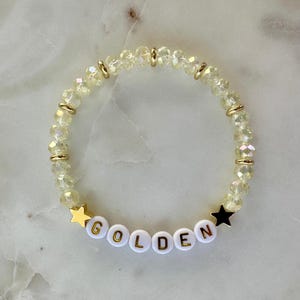 May include: A beaded bracelet with the word "GOLDEN" spelled out in white letter beads. The bracelet features gold-colored accents and star-shaped charms. The beads are a light yellow color.