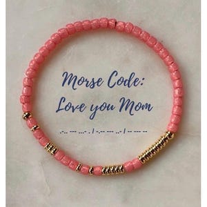 May include: A circular beaded bracelet featuring small, textured coral-pink beads interspersed with polished gold-tone metallic beads. The text "Morse Code: Love you Mom" is centered within the bracelet, accompanied by the corresponding Morse code dots and dashes.