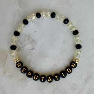 May include: A beaded bracelet with the word "REPUTATION" spelled out in gold letters on black beads. The bracelet also features clear and black beads, with gold accents.