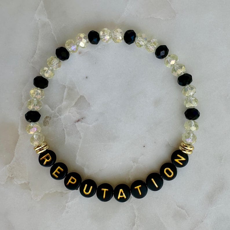 May include: A beaded bracelet with the word "REPUTATION" spelled out in gold letters on black beads. The bracelet also features clear and black beads, with gold accents.