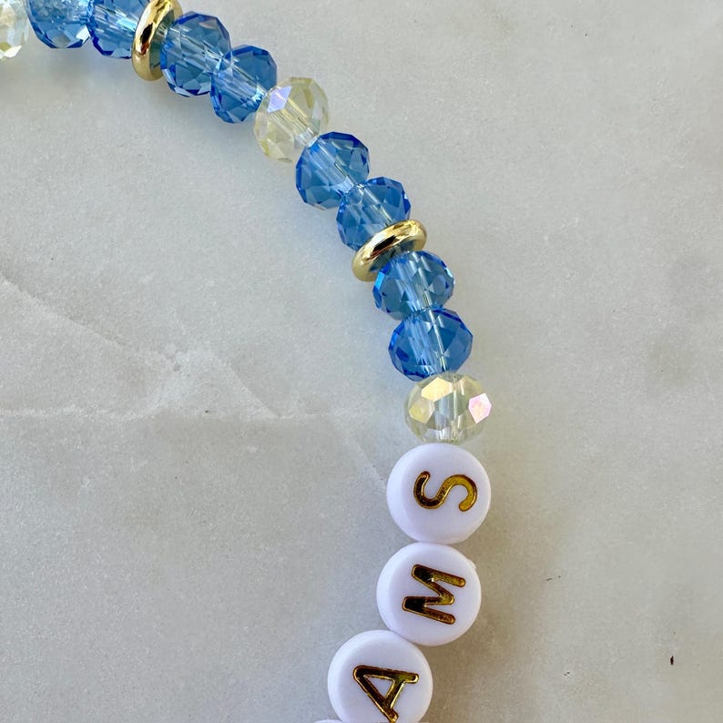 May include: A beaded bracelet with blue and clear crystal beads, gold-coloured spacers, and white letter beads spelling "AMA". The bracelet is strung on a flexible cord.