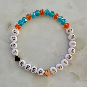 May include: A beaded bracelet with the words "JUST KEEP SWIMMING" spelled out in white letter beads. The bracelet features orange and blue crystal beads and two heart-shaped beads, one black and one copper.