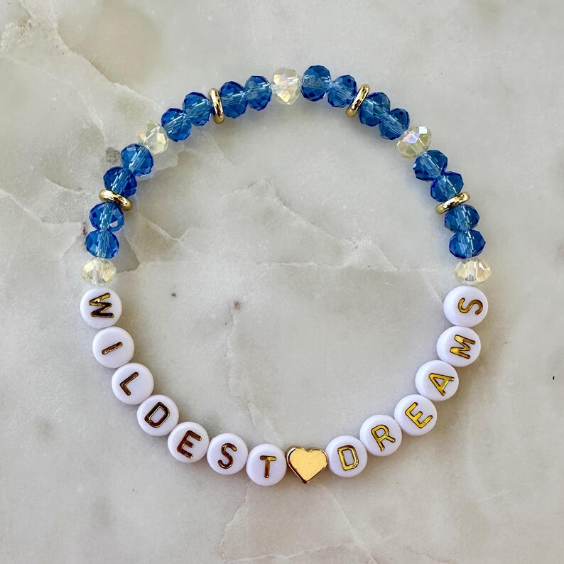 May include: A bracelet made of white beads with gold lettering spelling "WILDEST DREAMS". The bracelet also features blue and clear crystal beads, gold spacers, and a gold heart charm.