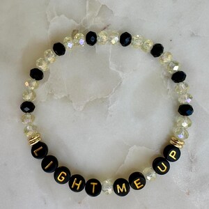 May include: A beaded bracelet with the words "LIGHT ME UP" spelled out in gold letters on black beads. The bracelet also features clear and black beads, and gold accents.