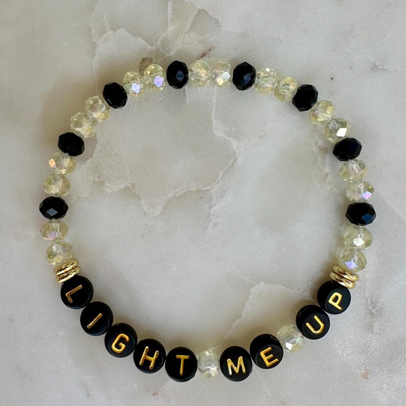 May include: A beaded bracelet with the words "LIGHT ME UP" spelled out in gold letters on black beads. The bracelet also features clear and black beads, and gold accents.