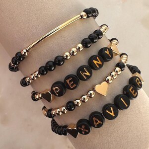 May include: Stack of personalized bracelets with black beads, gold beads, and gold-colored letter beads spelling out names. Some bracelets have gold heart charms and a gold bar.