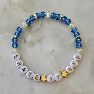 May include: A beaded bracelet with blue, clear, and gold beads. The bracelet features white letter beads spelling "SHAKE IT OFF" with gold star accents. The bracelet is designed to be worn on the wrist.