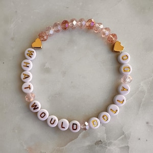 May include: A beaded bracelet featuring white round beads with gold lettering that spells out WHAT WOULD DOLLY DO. The design includes faceted light pink crystal beads and two gold-toned heart-shaped charms positioned symmetrically on the bracelet.