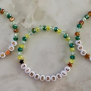 May include: Three beaded bracelets. One spells "NEVER GROW UP", another "TINKERBELL", and the third "YOU CAN FLY". They have gold heart and star charms, and are strung with green, gold, and clear beads.