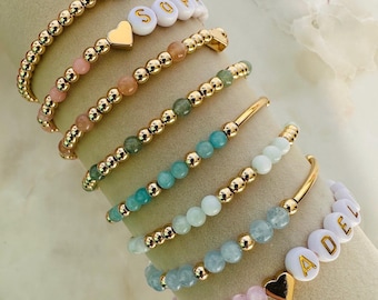Può includere: Una collezione di braccialetti dorati con perline di vari colori e perline con lettere bianche che compongono le parole "MOM" e "SOPHIA". Alcuni braccialetti presentano ciondoli a forma di cuore. I braccialetti sono esposti su una superficie neutra.