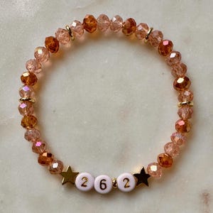 May include: A beaded bracelet with a mix of peach and amber-colored faceted beads. The bracelet includes gold-colored spacers and star-shaped charms. The word "262" is spelled out with white letter beads.