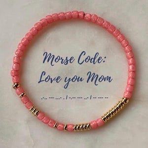 Könnte beinhalten: Rosa Perlenarmband mit goldenen Akzenten. Das Armband trägt den Text "Morse Code: Love you Mom" und die entsprechenden Morsezeichen.