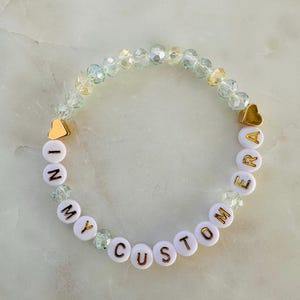 May include: A bracelet with the words "IN MY CUSTOM ERA" in gold lettering on white beads. The bracelet includes gold heart-shaped details and clear, iridescent beads. The bracelet is a fashionable accessory.