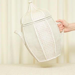 May include: A large, white, woven teapot with a domed lid and a curved spout. The teapot has a lattice-like pattern and is held by a hand. The background is a cream-colored curtain.