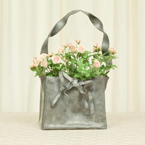 以下が含まれることがあります： ピンクのバラと緑の葉で満たされた灰色の金属製トートバッグ。 バッグには、お揃いの灰色のリボンが蝶結びで結ばれており、ねじれたハンドルが付いています。 花はバッグの中に配置され、花のディスプレイを作成しています。