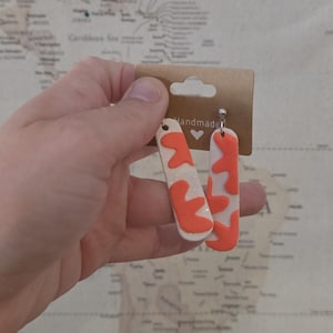 May include: Handmade earrings with a white base and orange abstract shapes. The earrings are rectangular and dangle from a silver hook. The earrings are displayed on a brown card with the word "handmade" printed on it.