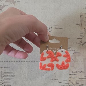 May include: Handmade earrings with a white base and coral-colored abstract shapes. The earrings are held up by a brown tag with the word "Handmade" printed on it. The earrings are on a map background.