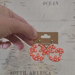 May include: Handmade earrings with a unique design. The earrings feature a white base with an orange, dotted pattern. The earrings are held by a person in front of a world map background. The earrings are attached to a brown card.