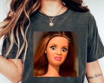 May include: Dark gray t-shirt featuring a square image of a doll with a surprised expression. The doll has red hair, large eyes, and a pursed mouth. The model is wearing blue jeans and white sneakers.