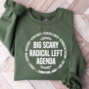 May include: An olive green sweatshirt with the text "BIG SCARY RADICAL LEFT AGENDA" in a white circular design. The design includes the phrases "VOTING RIGHTS", "CLEAN AIR & WATER", "REPRODUCTIVE FREEDOM", and "LOVE WINS".