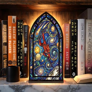 Puede incluir: Un panel de vidrieras que representa a un astronauta con un traje espacial sobre un fondo de galaxia arremolinada. El panel está iluminado desde atrás y se encuentra en un estante con libros y una taza negra. El panel mide unos 30 cm de alto.