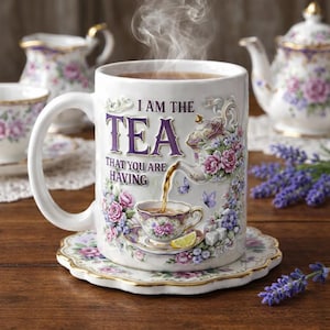 May include: White ceramic mug with the text "I AM THE TEA THAT YOU ARE HAVING" and floral designs. The mug is on a matching saucer, with a teapot and other teacups in the background. Steam rises from the mug.