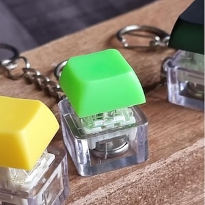 以下が含まれることがあります： カラフルなメカニカルキーボードのキーキャップが付いたキーホルダーのクローズアップ。キーキャップは正方形で、黄色、緑、黒などの明るい色があります。各キーキャップは、透明な長方形のスイッチハウジングに取り付けられています。