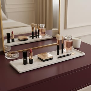 May include: A dressing table arrangement featuring cosmetics and accessories. Includes lipsticks, makeup brushes, a compact, perfume, and a pen on a marble tray. A mirror reflects the items, with a bed in the background. The color scheme is neutral with gold accents.