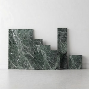 May include: A collection of green marble blocks of varying sizes and heights. The marble features a natural veining pattern in white and gray, creating a textured appearance. The blocks are arranged in a stepped configuration against a white wall.