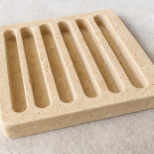 Puede incluir: Un jabonera cuadrada beige con seis ranuras paralelas y curvas. La jabonera est&aacute; hecha de un material de piedra texturizada y est&aacute; dise&ntilde;ada para sostener una pastilla de jab&oacute;n. El color neutro y el dise&ntilde;o sencillo la hacen adecuada para cualquier ba&ntilde;o.