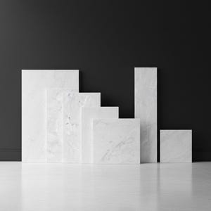 Puede incluir: Una colección de losas de mármol blanco de varios tamaños y formas, dispuestas en una exhibición escalonada. Las losas tienen sutiles vetas grises y están colocadas contra una pared gris oscuro y un suelo blanco.
