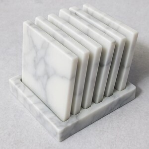 Puede incluir: Un juego de seis posavasos cuadrados de m&aacute;rmol con un veteado gris, expuestos en un soporte rectangular a juego. Los posavasos est&aacute;n hechos de m&aacute;rmol blanco pulido.