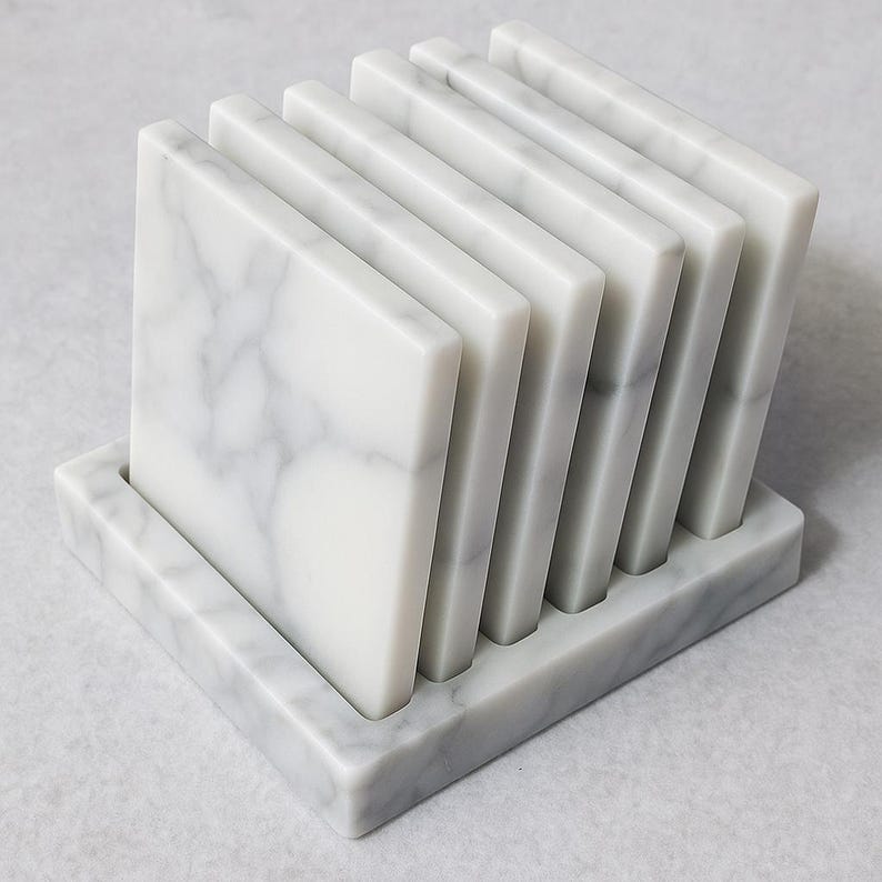 Puede incluir: Un juego de seis posavasos cuadrados de m&aacute;rmol con un veteado gris, expuestos en un soporte rectangular a juego. Los posavasos est&aacute;n hechos de m&aacute;rmol blanco pulido.