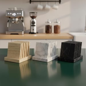 Può includere: Tre sottobicchieri quadrati in un ambiente cucina. Uno è in legno chiaro, uno in marmo bianco e il terzo in marmo nero. Ogni sottobicchiere ha una base con fessure verticali per contenere i sottobicchieri. Una macchina da caffè e un macinacaffè sono sullo sfondo.