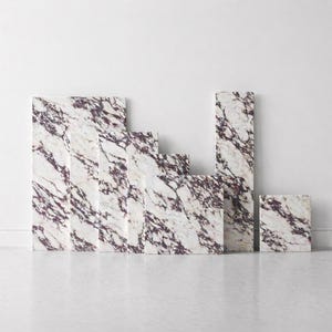 Puede incluir: Colección de baldosas de mármol de varios tamaños, dispuestas para crear una exhibición visual. Las baldosas presentan una base blanca con vetas moradas oscuras, creando una estética de piedra natural. La disposición está contra una pared y un suelo blancos.