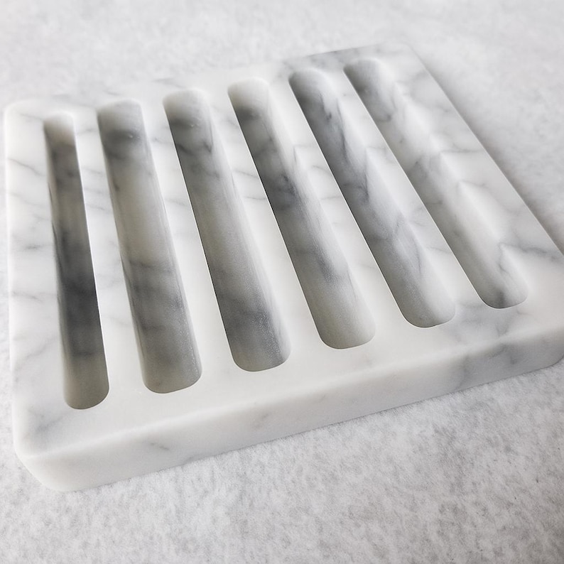Puede incluir: Un jabonera rectangular de m&aacute;rmol blanco con vetas grises. La jabonera tiene seis ranuras paralelas para el drenaje. Hecha de piedra natural, con un acabado liso y pulido.