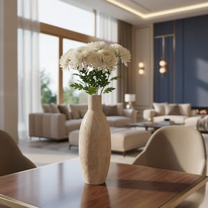 Op de afbeelding: Een beige vaas met witte bloemen en groene bladeren staat op een donkere houten tafel. De vaas staat in een moderne woonkamer met beige meubels, een blauwe accentmuur en grote ramen met transparante gordijnen. De kamer heeft een neutraal kleurenpalet.