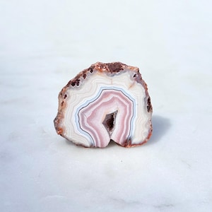 May include: A cross-section of a polished agate stone, showcasing concentric bands of color. The stone features a central cavity and a spectrum of colors, including white, pink, and brown. The stone is approximately 3 inches in diameter.