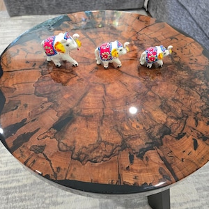 Puede incluir: Una mesa de centro redonda con un patrón de vetas de madera oscura y un borde de resina negro. Tres pequeñas figuras de elefantes blancos con mantas estampadas de colores están sobre la mesa.