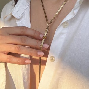 Può includere: Una collana color oro con un design a lariat unico. La collana presenta una delicata catena che forma un anello e si estende in una lunga e sottile goccia. La collana è indossata sopra una camicia bianca abbottonata.