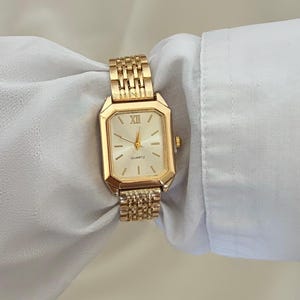 May include: A gold-toned rectangular wristwatch with a metallic link bracelet. The watch face features a light-coloured dial with gold-toned hour markers, a Roman numeral at the twelve o'clock position, and the word Quartz printed below the centre.