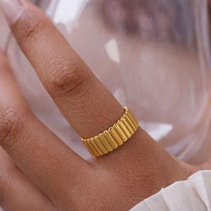 Könnte beinhalten: Ein goldfarbener Ring mit geripptem Design wird an einem Finger getragen. Die Oberfläche des Rings weist vertikale Linien auf, die ein strukturiertes Aussehen erzeugen. Der Ring ist der Fokus und zeigt ein schlichtes, aber elegantes Design.