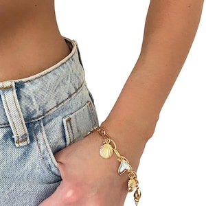 Könnte beinhalten: Goldfarbenes Charm-Armband mit Muschel-, Meerjungfrauenflossen- und Schneckenhaus-Anhängern. Das Armband wird am Handgelenk getragen, mit einer Jeans-Hosentasche im Hintergrund. Das Armband hat ein Kettenglied-Design.