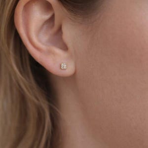 Könnte beinhalten: Nahaufnahme eines Ohrs mit einem kleinen, quadratischen Ohrstecker. Der Ohrring ist goldfarben und mit kleinen, funkelnden Steinen besetzt. Die Haut um das Ohr ist hell, und das Haar ist hellbraun.