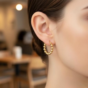 Könnte beinhalten: Goldene Creolen mit Perlenmuster. Die Ohrringe bestehen aus kleinen, runden, goldfarbenen Perlen, die zu einem Reifen aufgereiht sind. Die Ohrringe werden am Ohr getragen.