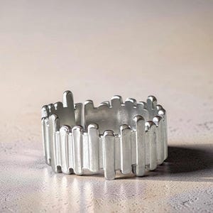 Könnte beinhalten: Ein silberfarbener Ring mit einem einzigartigen Design. Der Ring hat vertikale, rechteckige Stäbe, die das Band umschließen. Die Stäbe sind unterschiedlich hoch, wodurch eine unebene, strukturierte Oberfläche entsteht. Der Ring wird vor neutralem Hintergrund gezeigt.