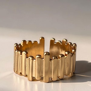 Könnte beinhalten: Goldfarbener Ring mit einem strukturierten Design aus vertikalen Stäben. Der Ring hat ein offenes Design für eine verstellbare Größe. Der glänzende, metallische Ring ist vor einem neutralen Hintergrund platziert.