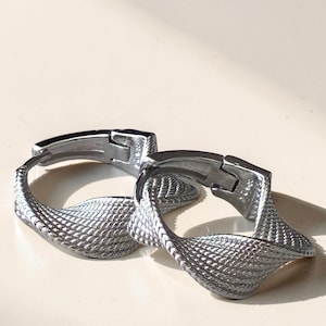 May include: A pair of silver-toned hoop earrings featuring a twisted, textured design with a cross-hatched pattern. The earrings have a hinged closure mechanism and a metallic finish, showcasing a continuous, flowing shape that curves into a loop.